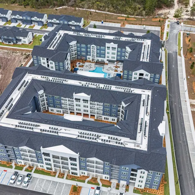 Aerial view of a large, modern apartment complex with multiple buildings and a central courtyard featuring a pool. Surrounding areas include under-construction sections and landscaping.