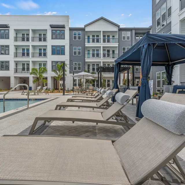 A relaxing poolside area with lounge chairs and a cabana, surrounded by palm trees and modern apartment buildings in the background.
