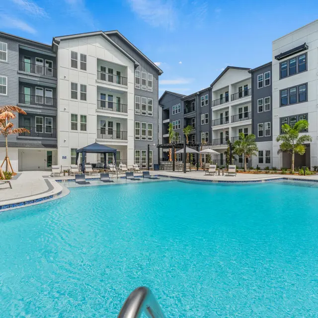 A spacious pool area surrounded by modern apartment buildings, featuring lounge chairs and palm trees under a clear blue sky.