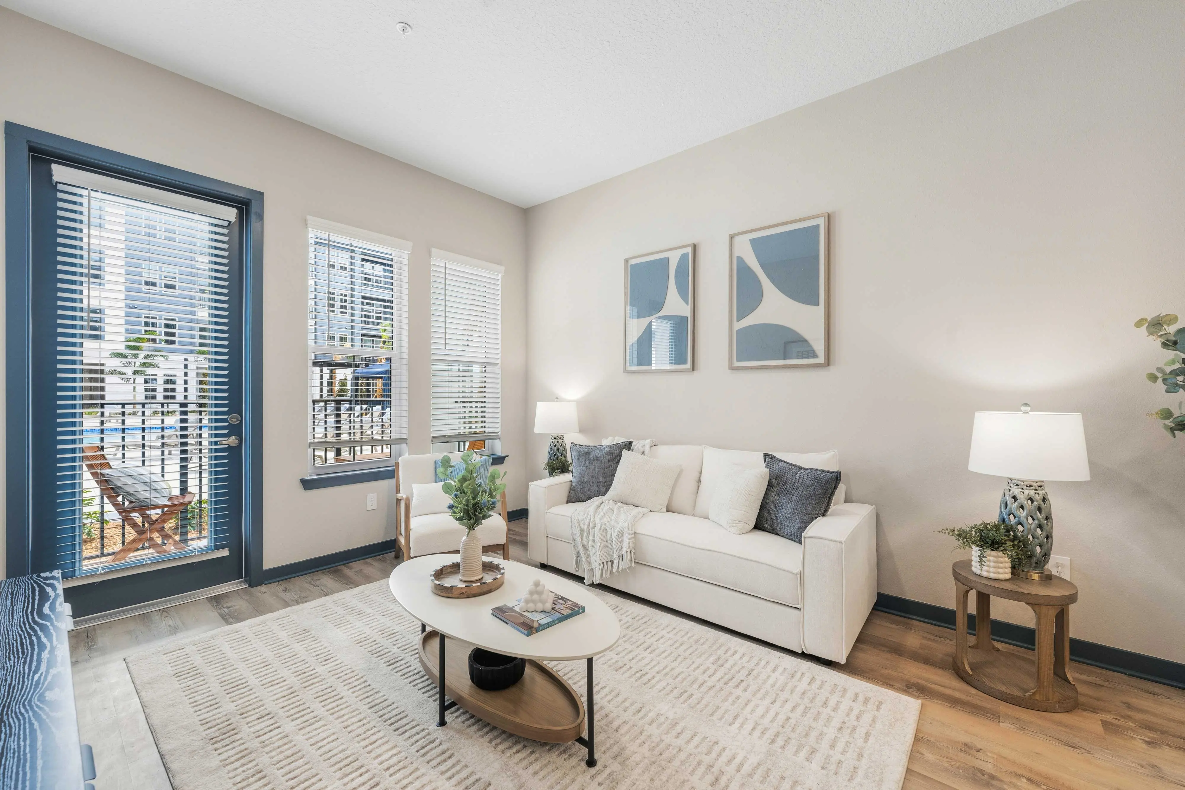 A modern living room featuring a white couch, two lamps, and a coffee table with decorative items. Large windows provide natural light, with a door leading to a balcony. Wall art is displayed above the couch.