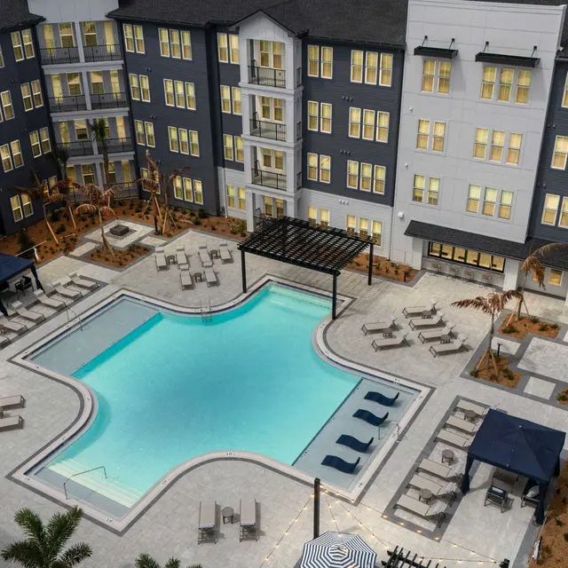 Aerial view of a modern apartment complex with a central pool area surrounded by lounge chairs and shaded cabanas.