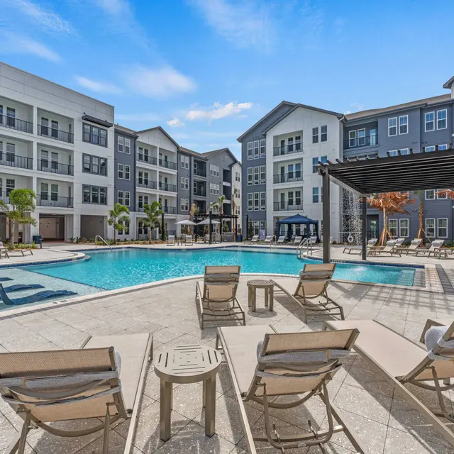 A modern apartment complex with a swimming pool in the center, surrounded by sun loungers and palm trees under a clear blue sky.