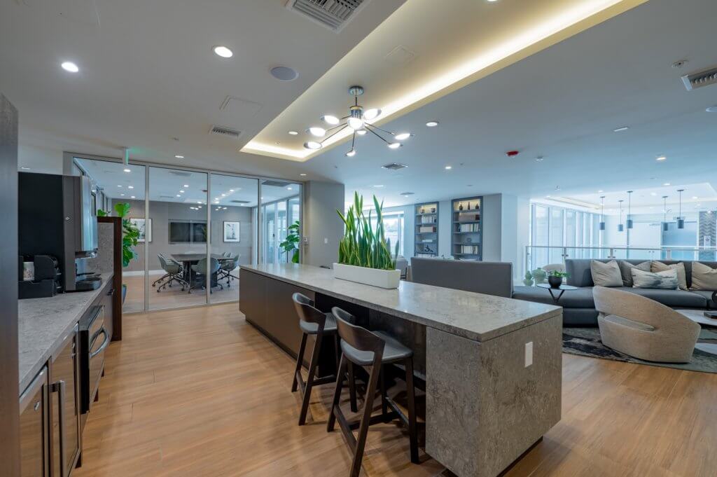 Modern kitchen and lounge area with a large countertop and seating, overlooking a conference room.