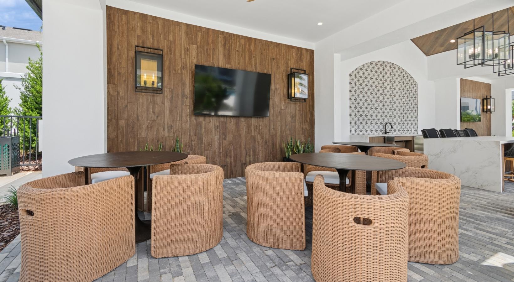 Pet-Friendly Luxury Apartments in Reunion, FL - Ascend Reunion Crossing - An Outdoor Lounge with Round Tables, Wicker Chairs, Framed Wall Sconces, and Mounted TV on a Wood-Paneled Wall.