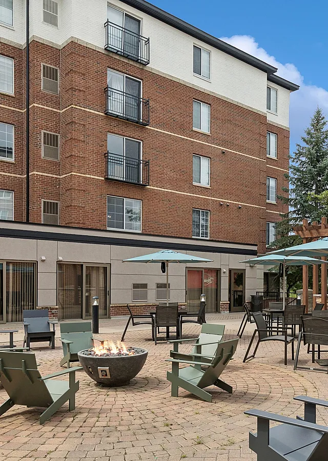 Charming outdoor courtyard with cozy fire pit, surrounded by Adirondack chairs and shaded dining tables, set against a modern apartment building facade.