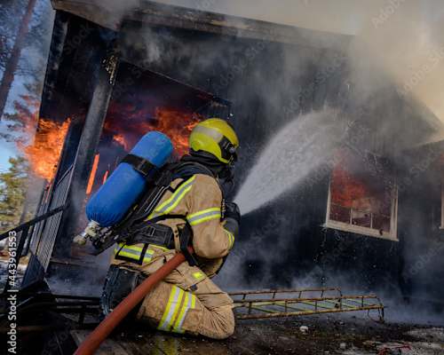 A firefighter in full gear is spraying water on a burning building
