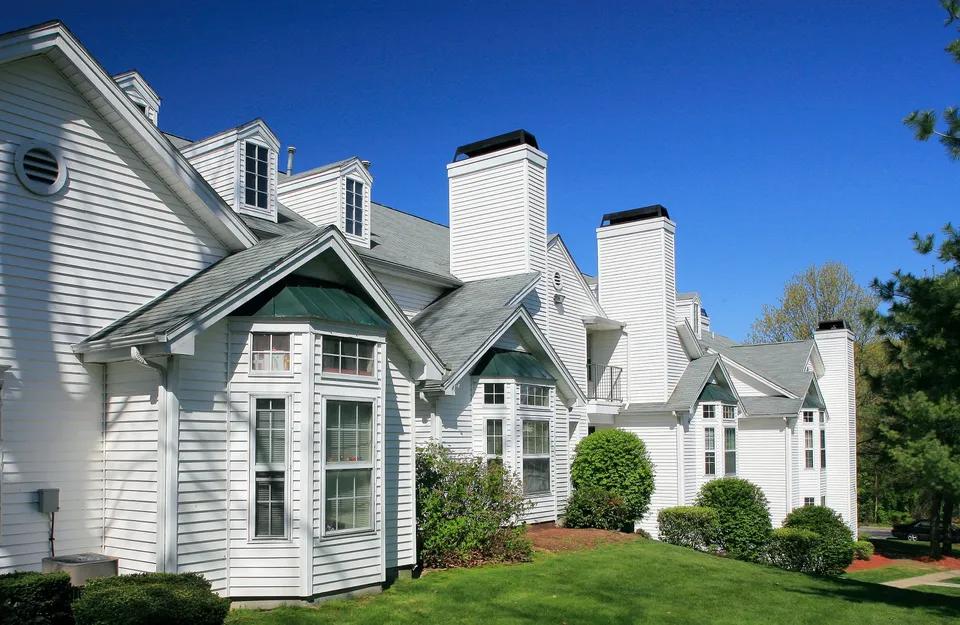 Charming white clapboard townhouse with elegant gabled roofs and large bay windows, surrounded by lush green landscaping, under a clear blue sky.