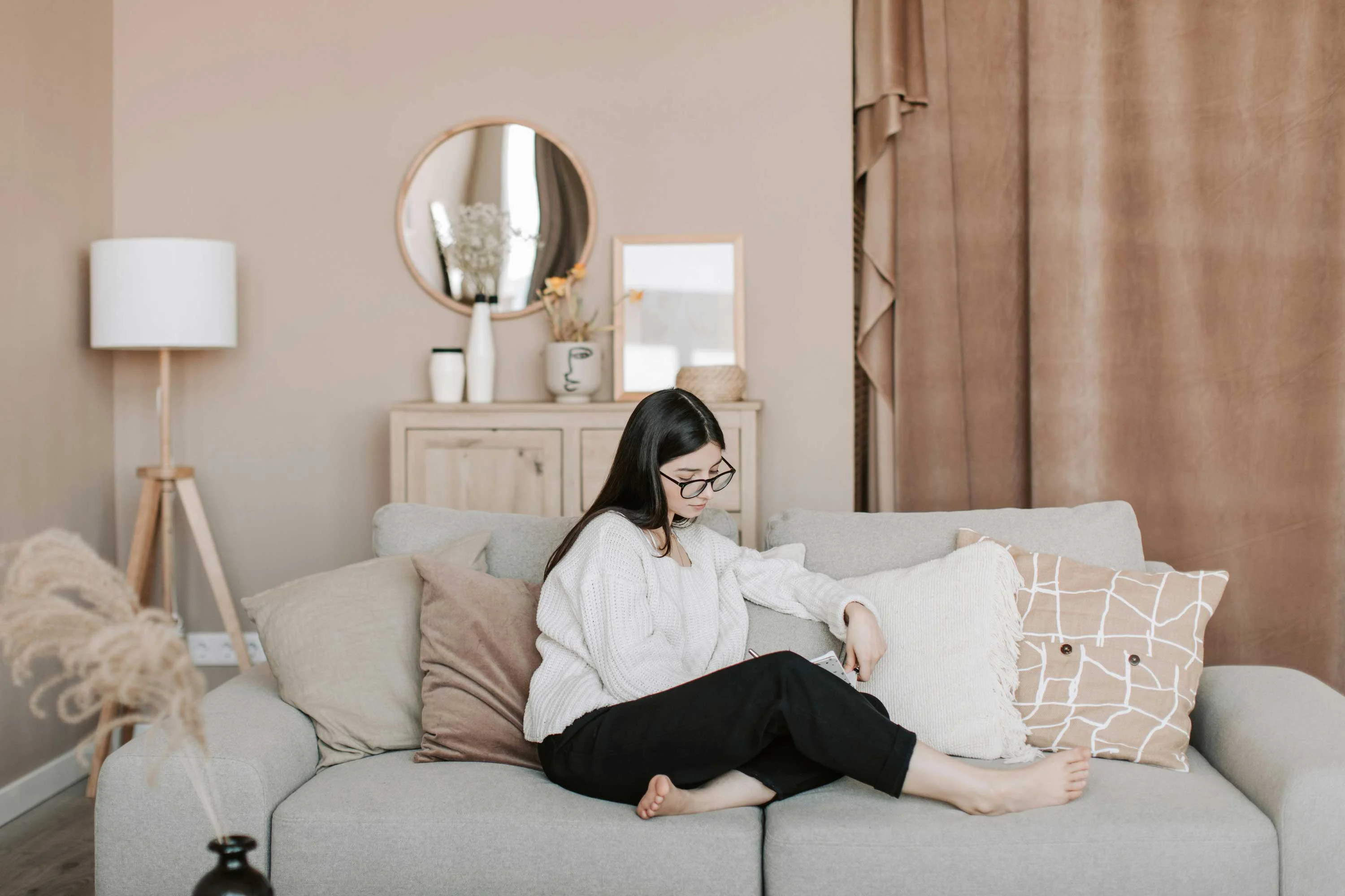 A young woman sitting on a couch with decorative pillows in a warm-toned living room. She is focused on something in her hands, wearing a casual outfit. The background features a round mirror, a lamp, and curtains.