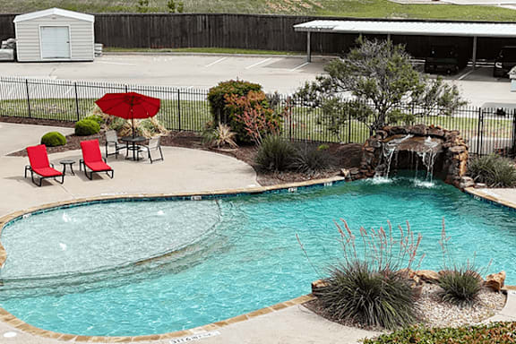 A pool with a waterfall and a patio with chairs and an umbrella.