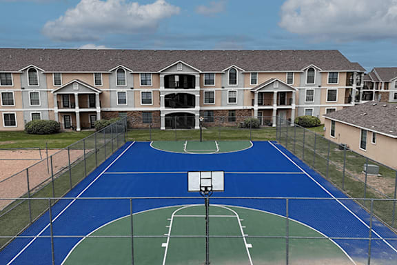 A basketball court is surrounded by a chain link fence and is located in front of apartment buildings.