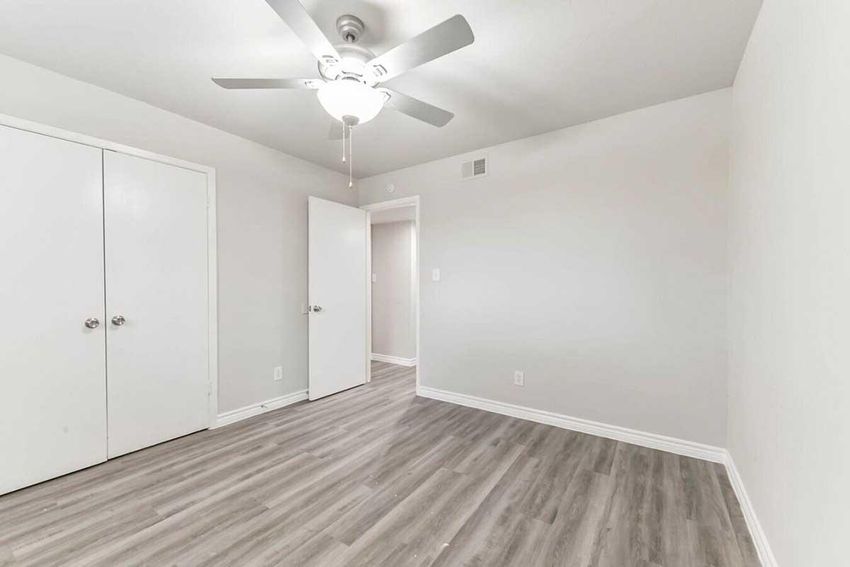 A clean, empty room featuring light-colored walls, a ceiling fan, and laminate flooring. Two closed white closet doors are on the left, with a partially opened door leading to another area. The space appears well-lit and tidy, ideal for various uses such as a bedroom or office.