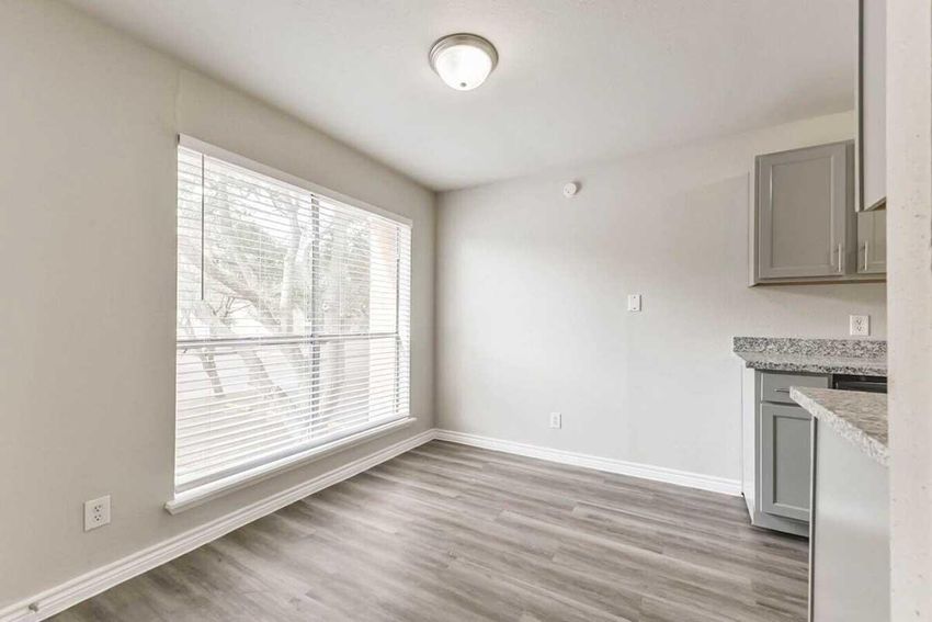 A bright and modern room featuring large windows with blinds, light gray walls, and wood-style flooring. There's a kitchen area with gray cabinets and a granite countertop, creating a clean and open space suitable for various uses.