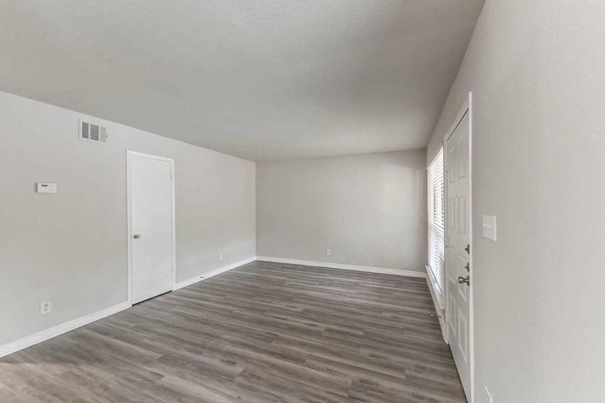 A spacious, empty room with light gray walls and a smooth wooden floor. A single door on the left leads to another area, while a white door on the right appears to be a closet. Natural light streams in from a window on the far wall, illuminating the uncluttered space.