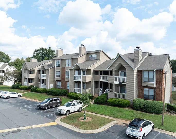 Charming apartment complex with modern architecture, ample parking, and lush greenery under a bright blue sky.