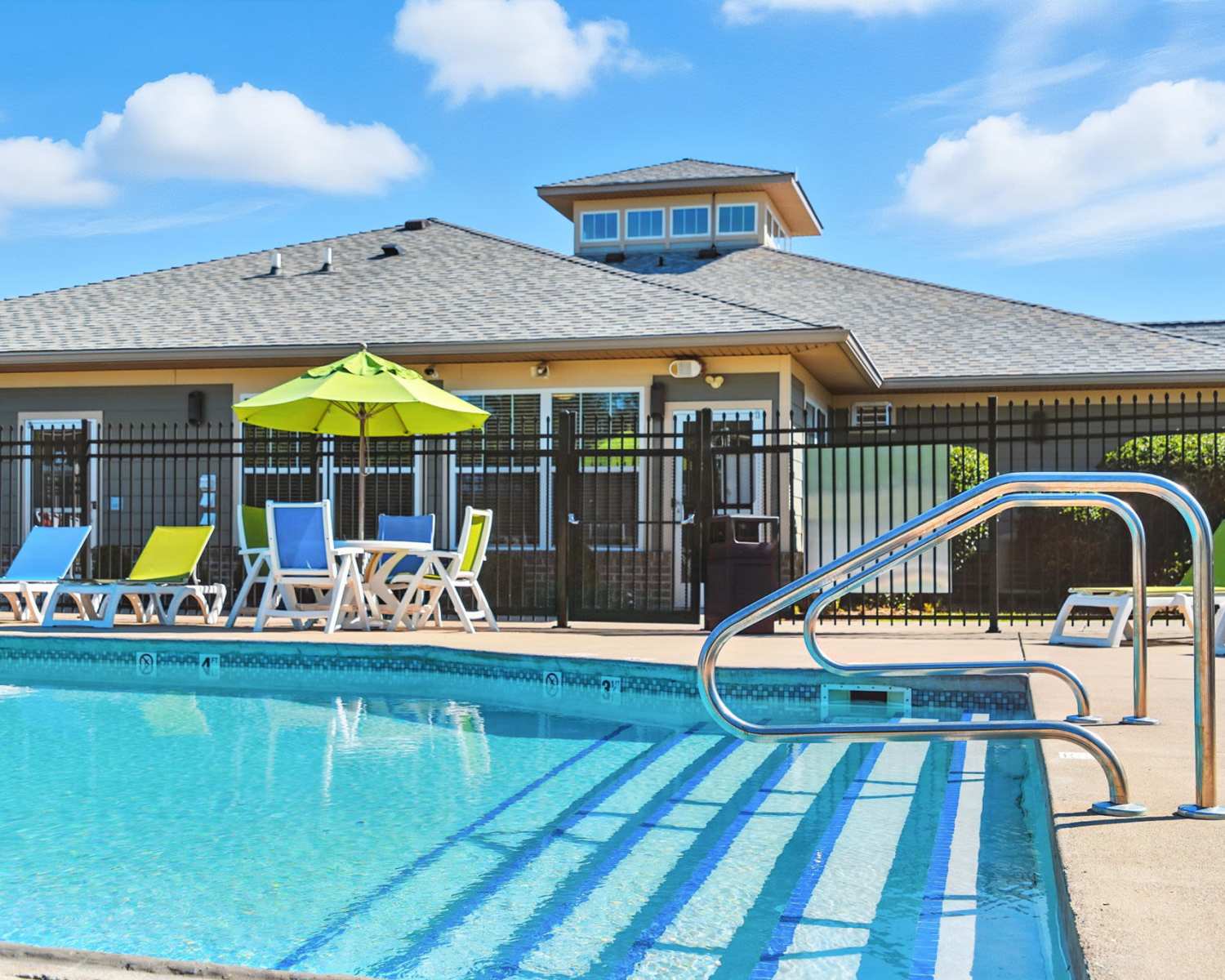 Sparkling pool with lounge chairs and green umbrella near a gated entrance to the community clubhouse at Bristol Bluffs Apartments in Louisville, Kentucky