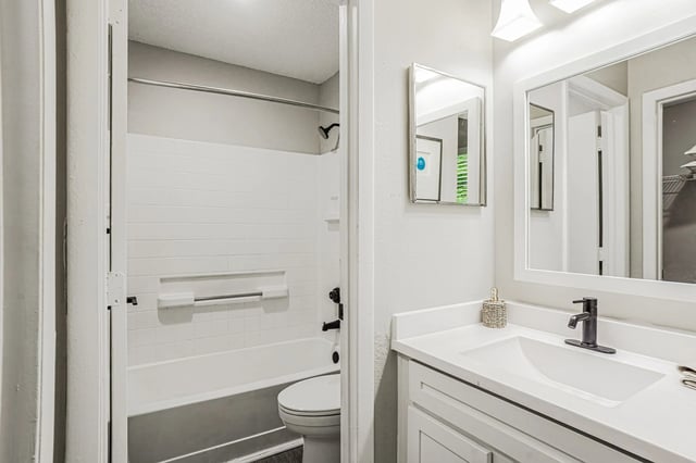 Modern bathroom with a white vanity, sink, and black faucet. Features a bathtub with white tile surround and a toilet.