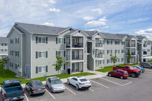 A parking lot in front of a grey apartment building with cars parked in the lot.