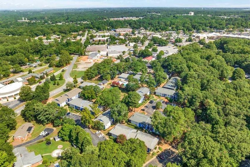 Aerial view of a suburban neighborhood surrounded by greenery, with residential buildings intermixed with trees. In the background, a commercial area is visible, featuring various shops and parking lots, showcasing a mix of urban and natural environments. Blue sky with scattered clouds is visible above.