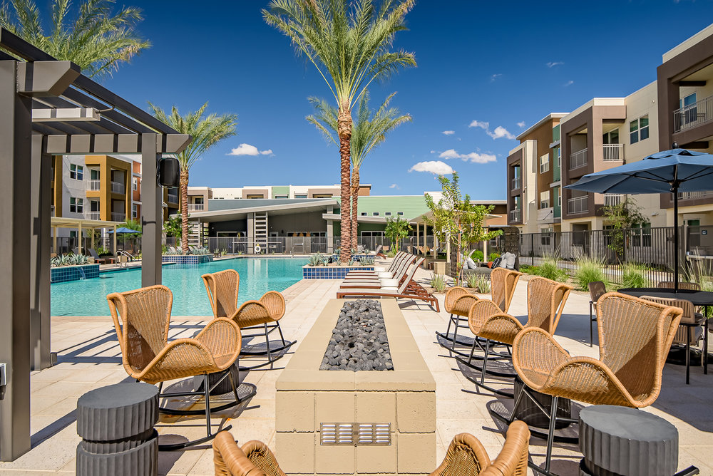 Outdoor pool area with wicker chairs, fire pit, palm trees, and modern apartment buildings in the background.