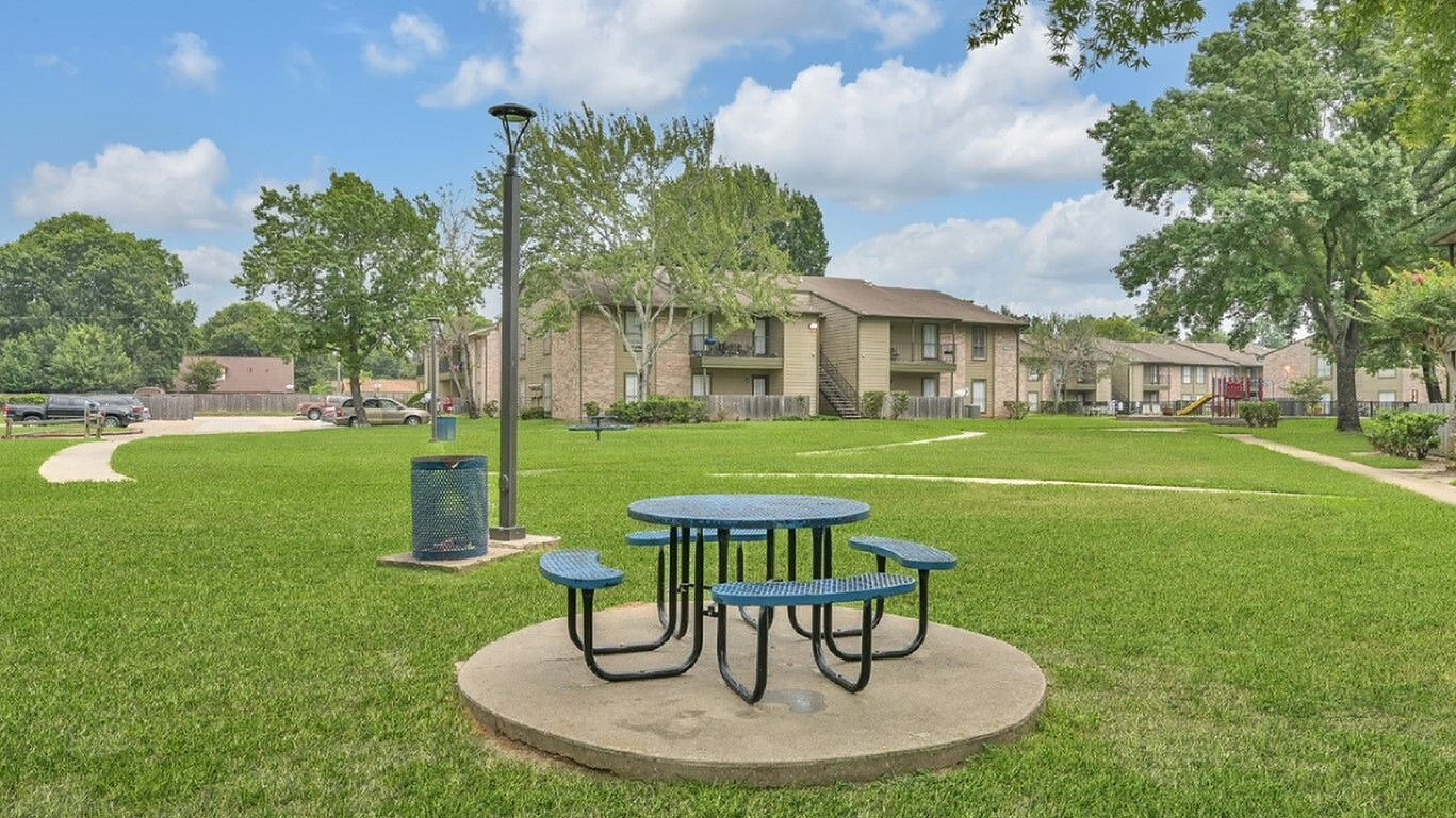 Inviting apartment complex with spacious green lawns, a cozy picnic area, and charming residential buildings under a vibrant blue sky.