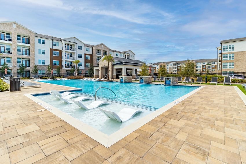 A bright and inviting outdoor pool area featuring lounge chairs along the edge, surrounded by landscaped greenery. The pool has a modern design with a hot tub and a shaded pavilion nearby. In the background, there are multiple apartment buildings with a mix of brick and siding facades under a clear blue sky.