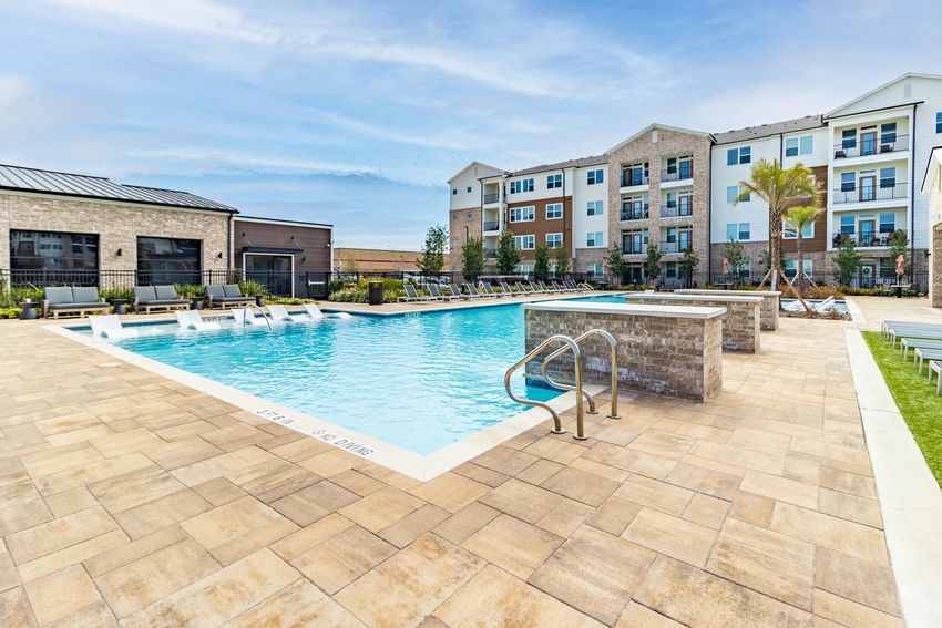 An outdoor swimming pool surrounded by lounge chairs and palm trees, located near a multi-story residential building. The pool area features a light-colored stone deck and clear blue water, with a few stairs leading into the pool. The sky is partly cloudy, indicating a pleasant day.