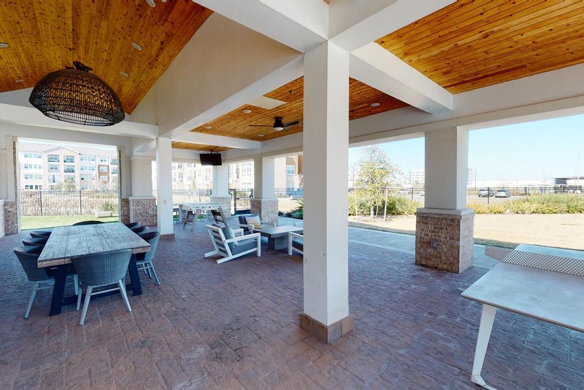 A spacious outdoor lounge area featuring a wooden ceiling, large windows, and a mix of dining tables and casual seating. There are stone pillars, pavers on the floor, and a view of a grassy area outside, creating a relaxed atmosphere for gatherings or relaxation.