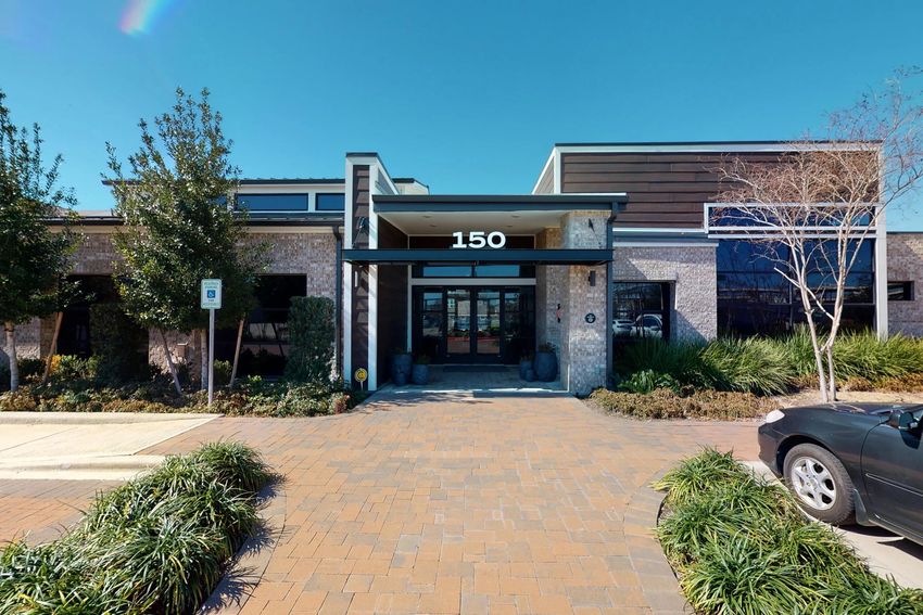 A modern building entrance featuring a large glass door, with the number "150" prominently displayed above. The façade combines brick and wood elements. In front, there's a landscaped area with shrubs and plants, along with a brick pathway and a parked car on the right. The sky is clear and blue.