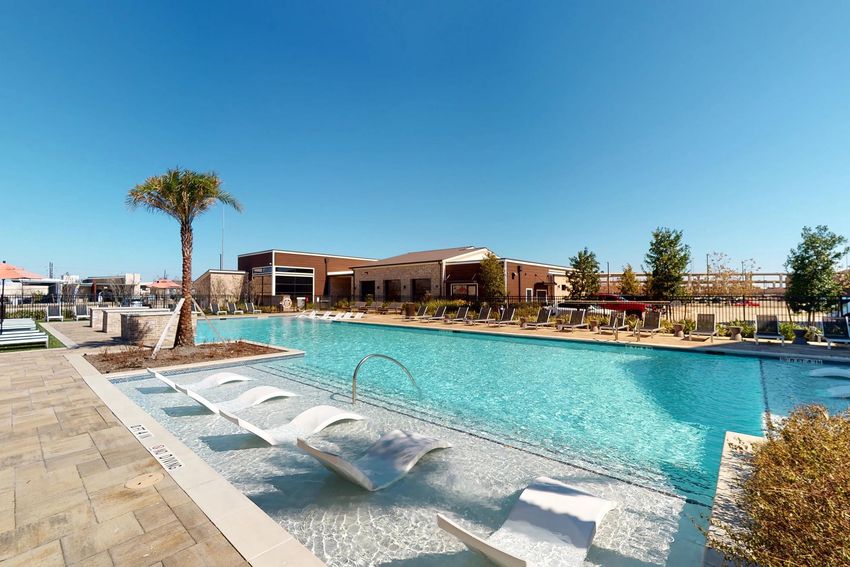 A spacious outdoor swimming pool surrounded by lounge chairs and palm trees, with a modern building in the background. The clear blue sky complements the serene atmosphere, making it an inviting space for relaxation and leisure.