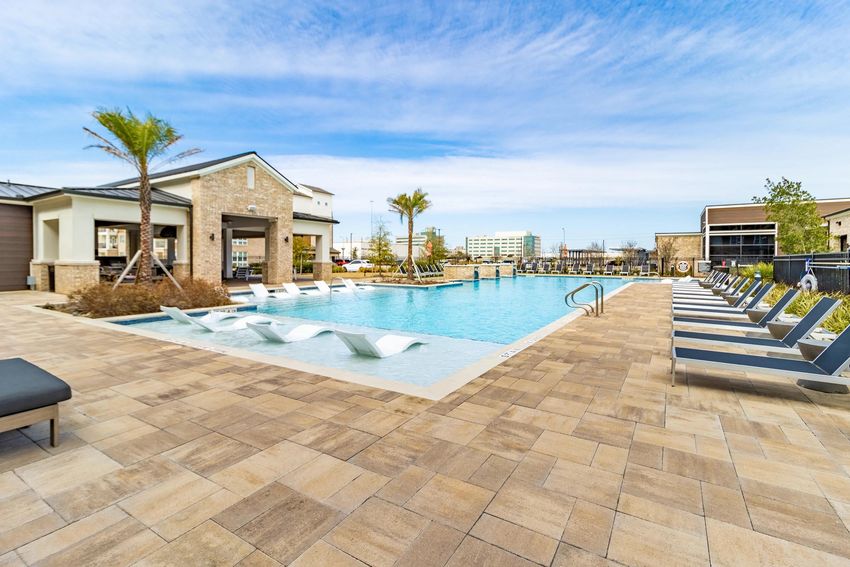 A modern outdoor swimming pool surrounded by lounge chairs, with palm trees in the background. The pool area features a sleek design, with a building nearby, and clear blue skies overhead, creating a tranquil, inviting atmosphere for relaxation.