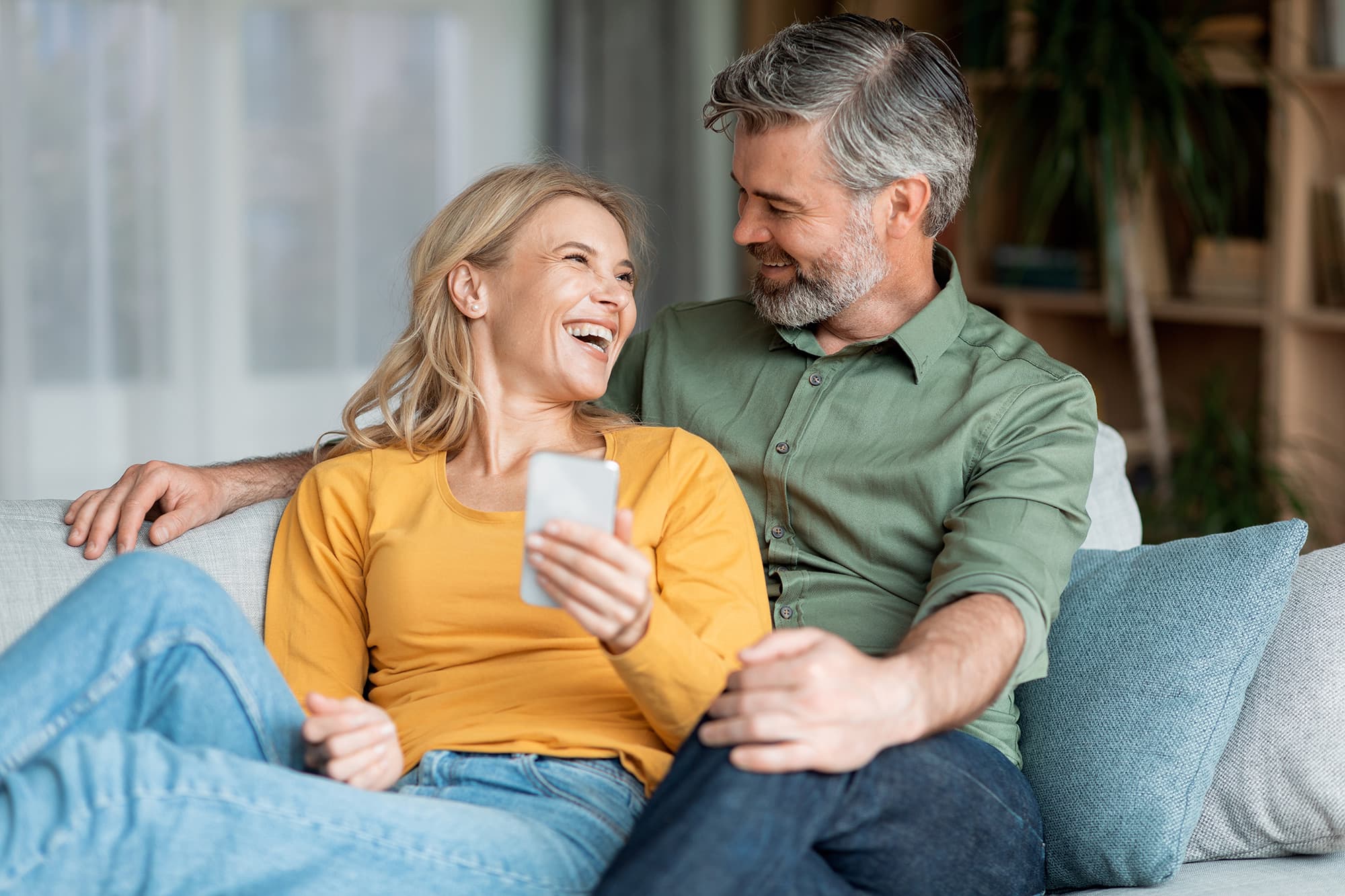A smiling couple relaxing together on a couch and looking at a smartphone at Livana Fair Lawn in New Jersey.