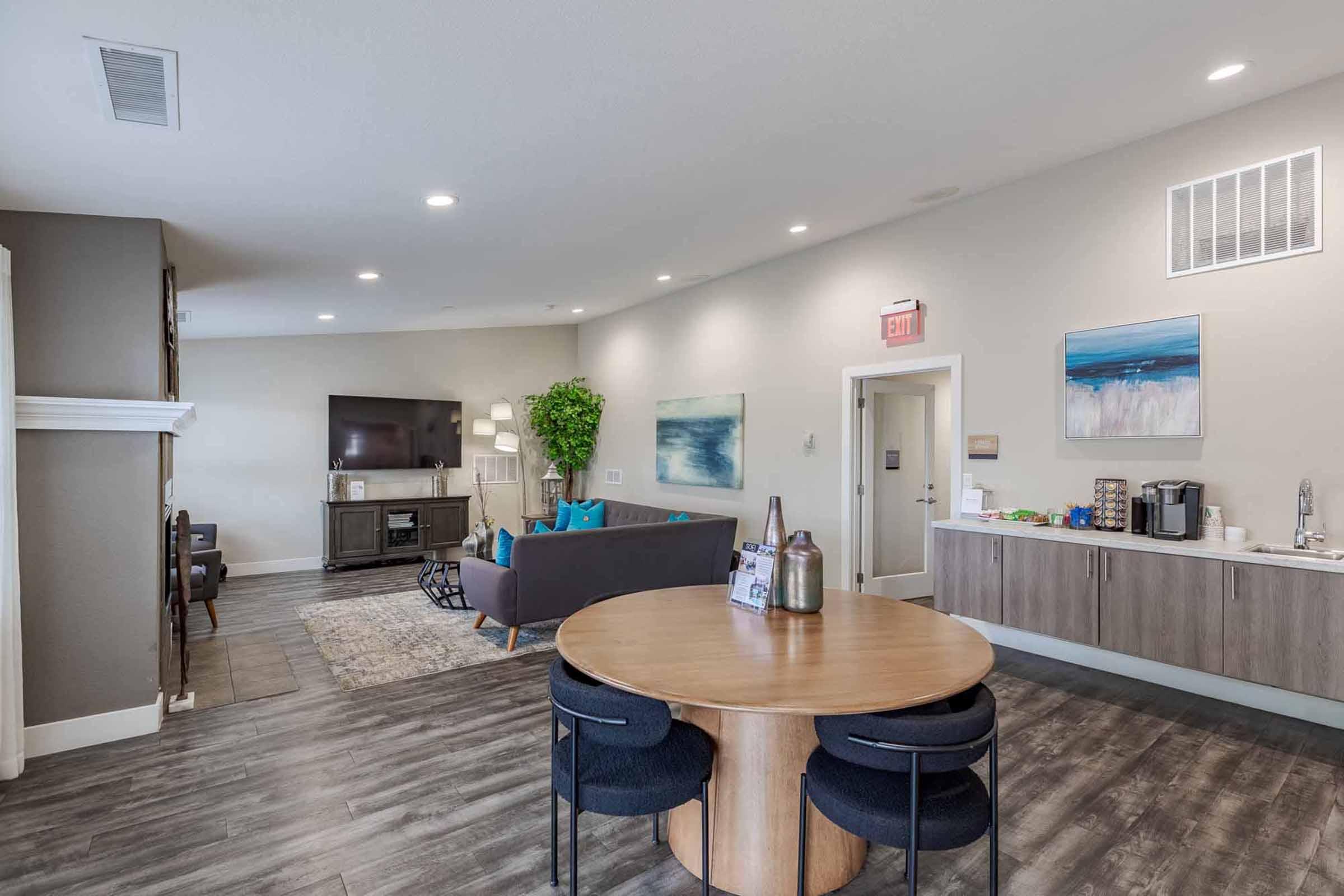 A modern lounge area with a gray sofa adorned with blue pillows, a round wooden dining table with black chairs, a television mounted on the wall, and artwork featuring abstract ocean scenes. A beverage station is visible on a cabinet, and a potted plant adds a touch of greenery to the space.