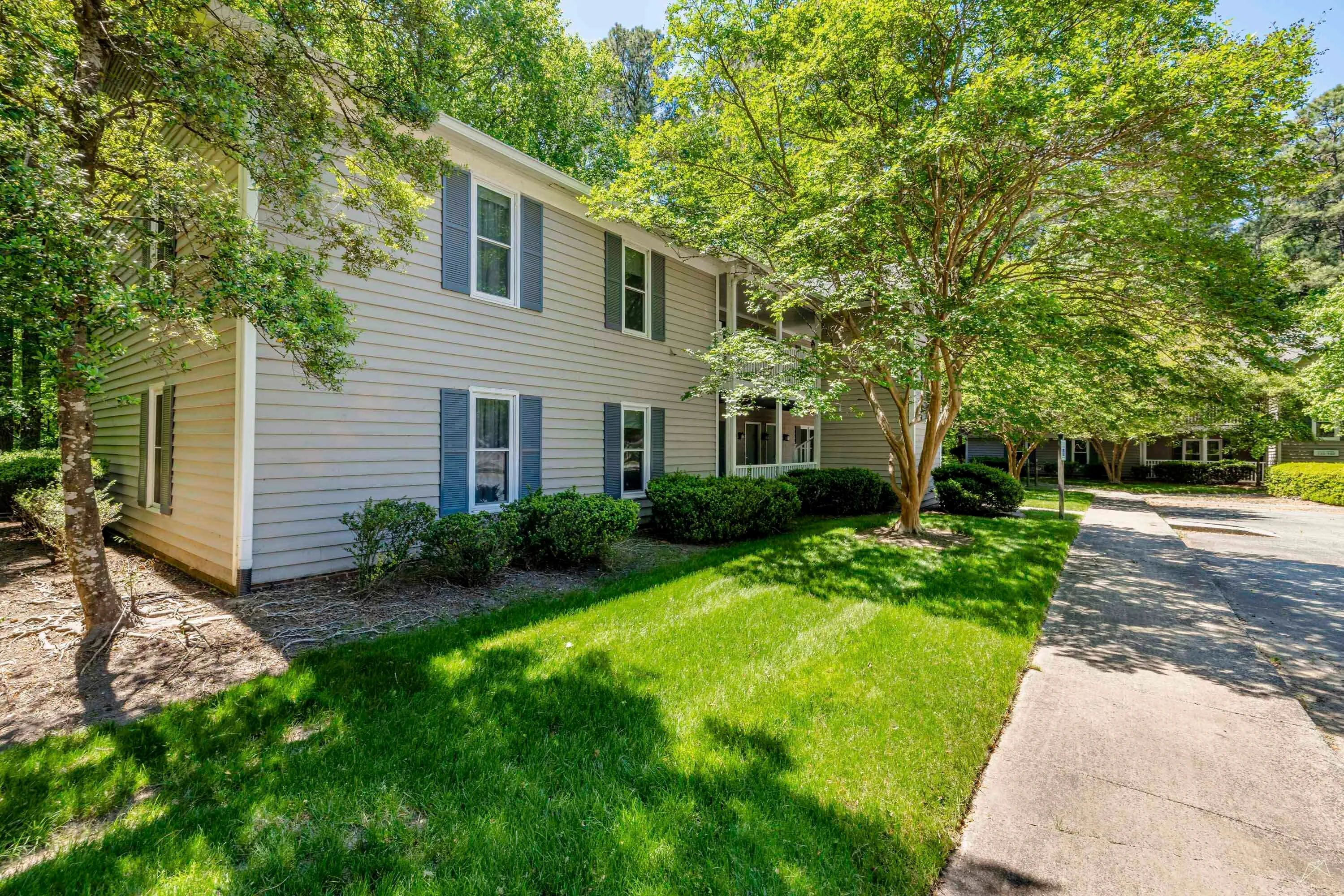 A well-maintained apartment complex with green lawns, shrubs, and a paved walkway surrounded by trees.