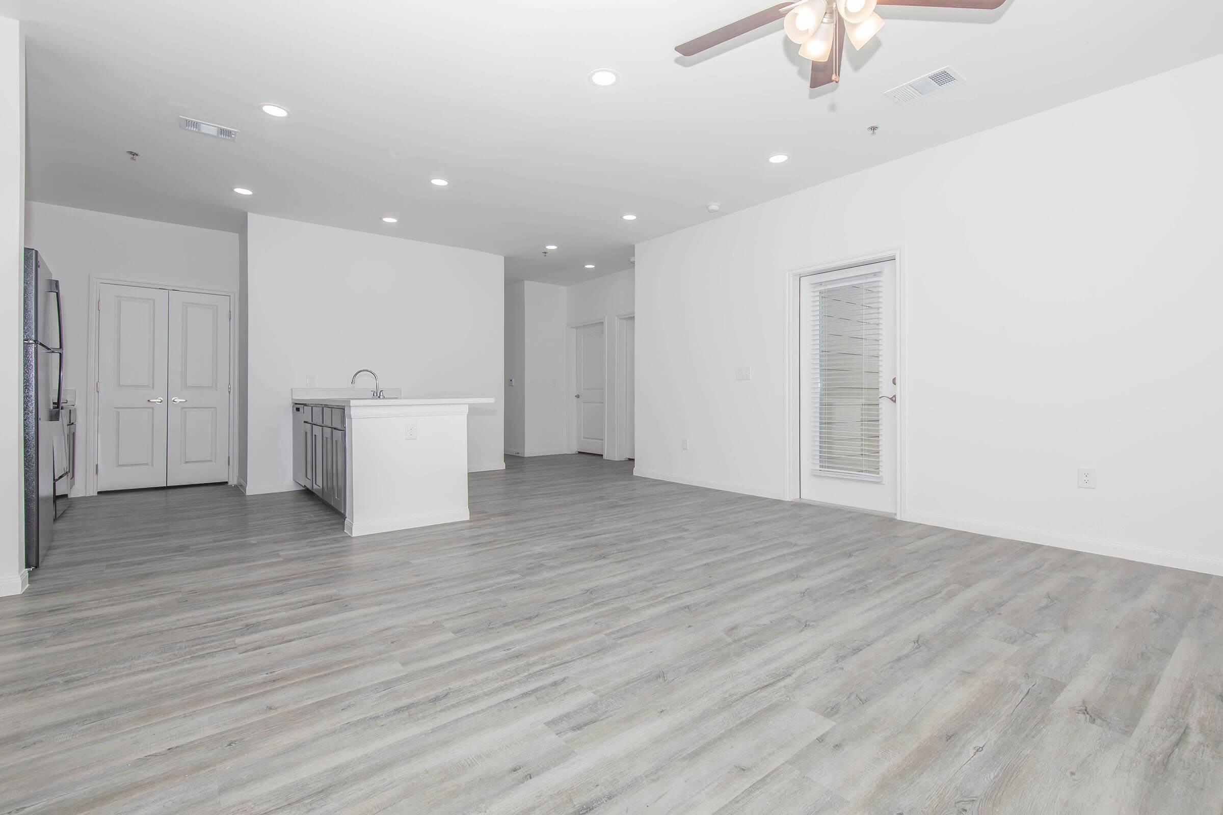 Spacious, modern living area with light gray wood-style flooring, a ceiling fan, and ample natural light. The kitchen features a large island with seating, white cabinetry, and stainless steel appliances. The walls are painted in a bright white, enhancing the open and airy feel of the space.