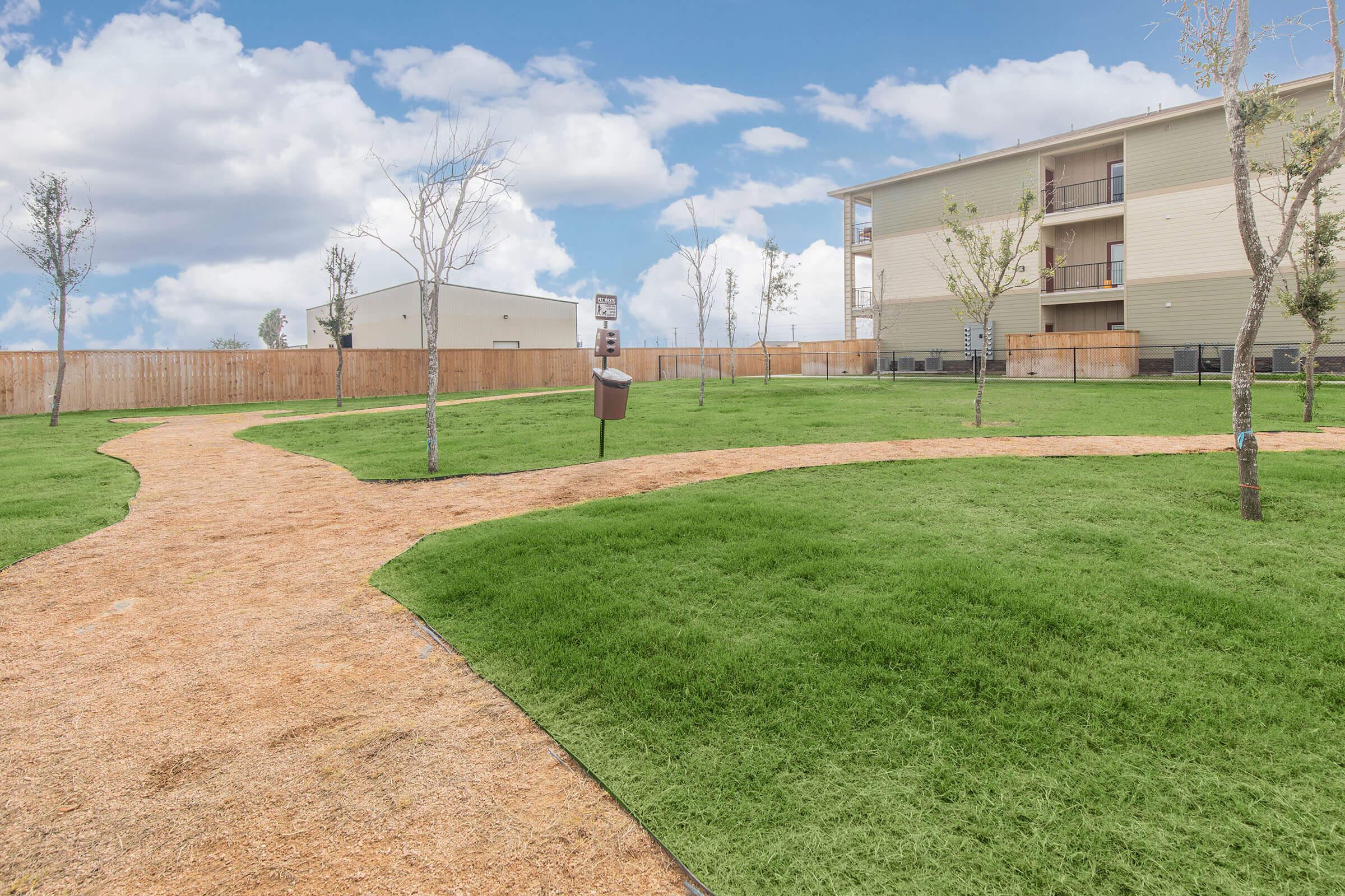 A landscaped outdoor area featuring a grassy path winding through freshly planted trees. In the background, a multi-story building and a wooden fence are visible under a partly cloudy blue sky.