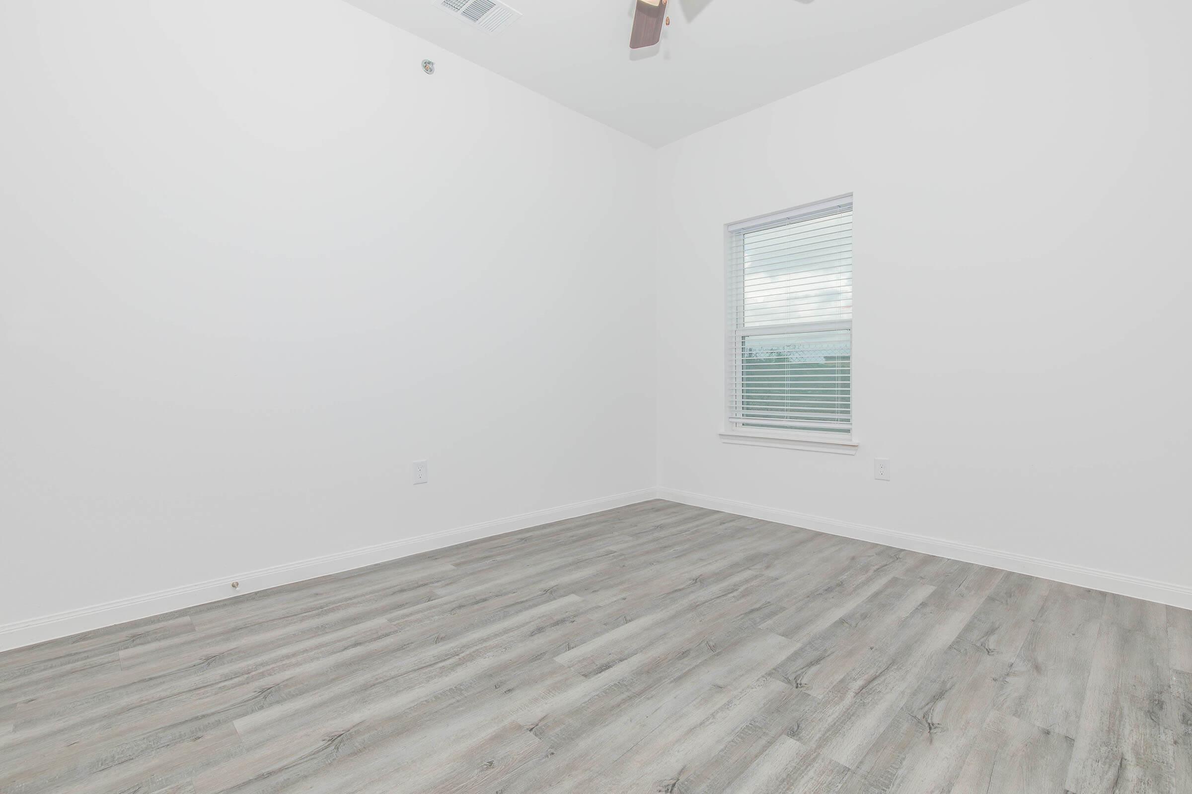 Interior of an empty room with light gray wooden flooring, white walls, and a small window with blinds. The ceiling features a ceiling fan, and the space appears well-lit and airy.