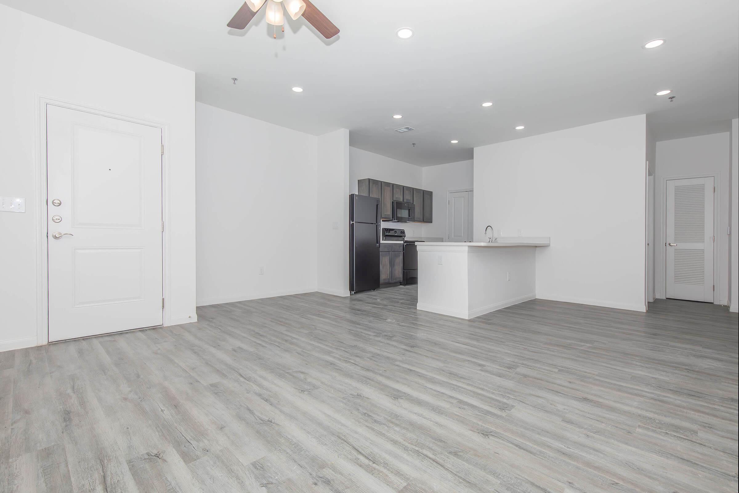 Spacious, modern living area with light wood-style flooring and a ceiling fan. The open layout features a kitchen with stainless steel appliances and a breakfast bar. A white door leads to the entrance. Natural light floods the space through recessed lighting, creating a bright and inviting atmosphere.