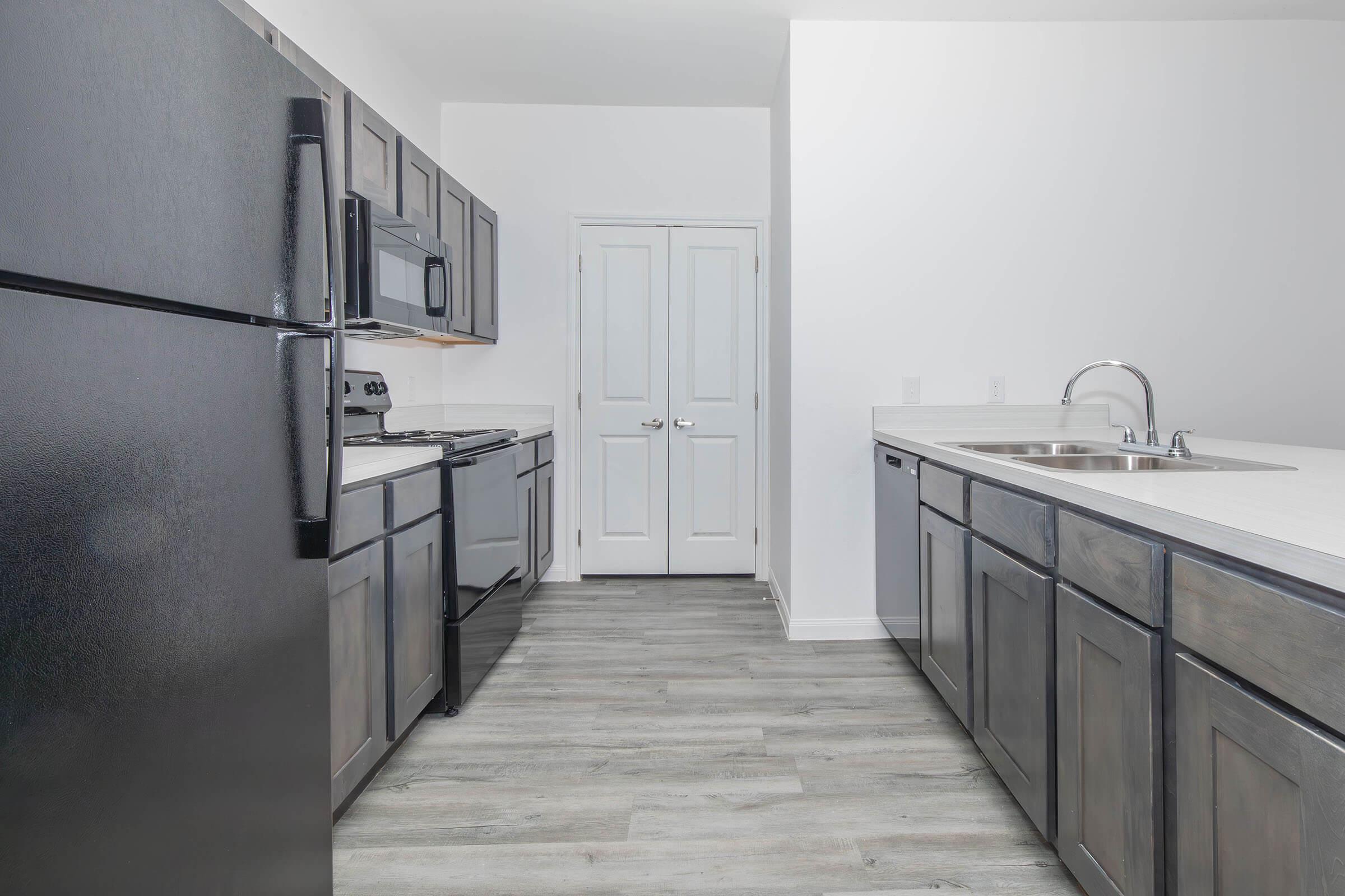 A modern kitchen featuring black cabinetry and appliances, including a refrigerator, oven, and microwave. The countertops are light-colored, enhancing the space. There is a double sink and a dishwasher. The flooring is wood-like, and the walls are painted white, providing a clean and bright atmosphere.