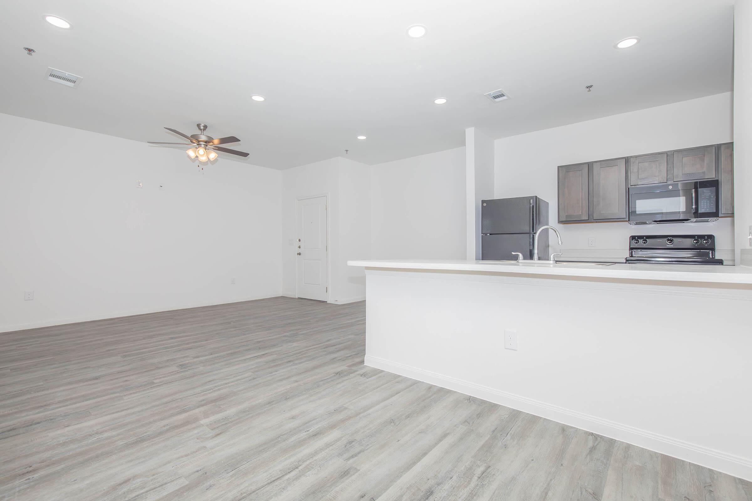 Modern, open-concept living space featuring light wooden flooring, white walls, and a ceiling fan. The kitchen includes dark cabinetry, a stainless steel refrigerator, and an electric stove. The area is well-lit with recessed lighting and has an inviting, spacious feel.