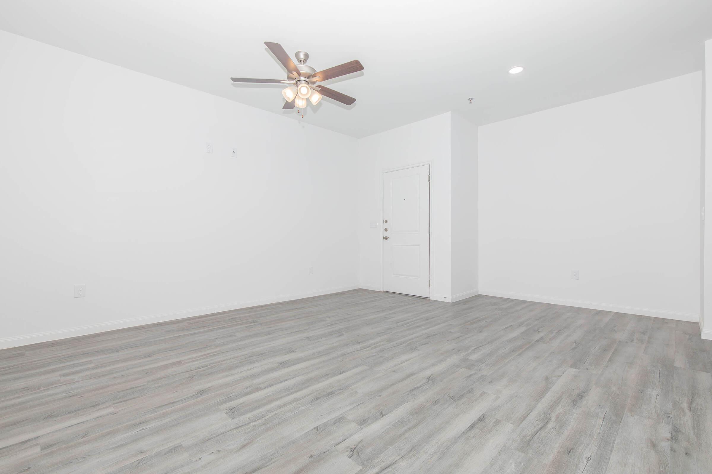 A spacious, empty room featuring light-colored walls and a wood-like floor. A ceiling fan with five light fixtures hangs from the ceiling. There is a white door leading outside, and the overall atmosphere is bright and minimalistic, providing a blank canvas for decoration.