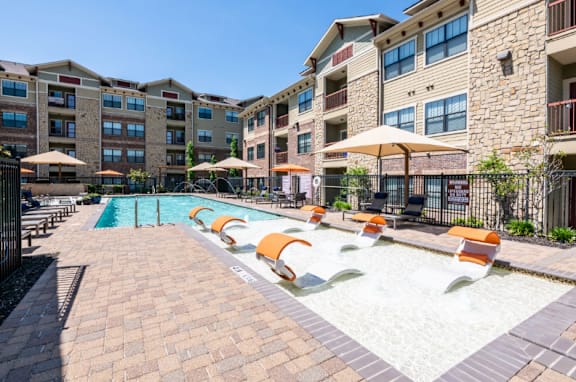 A swimming pool surrounded by orange lounge chairs and umbrellas in front of apartment buildings.