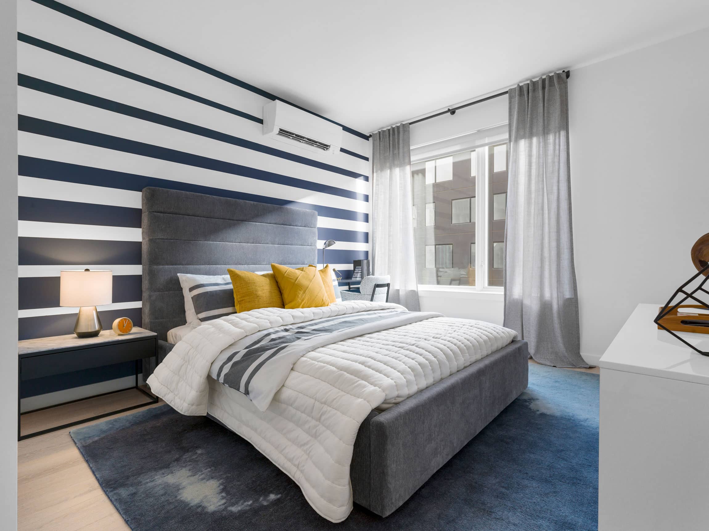 modern apartment bedroom with gray padded headboard, gold accent pillows, striped accent wall and large window