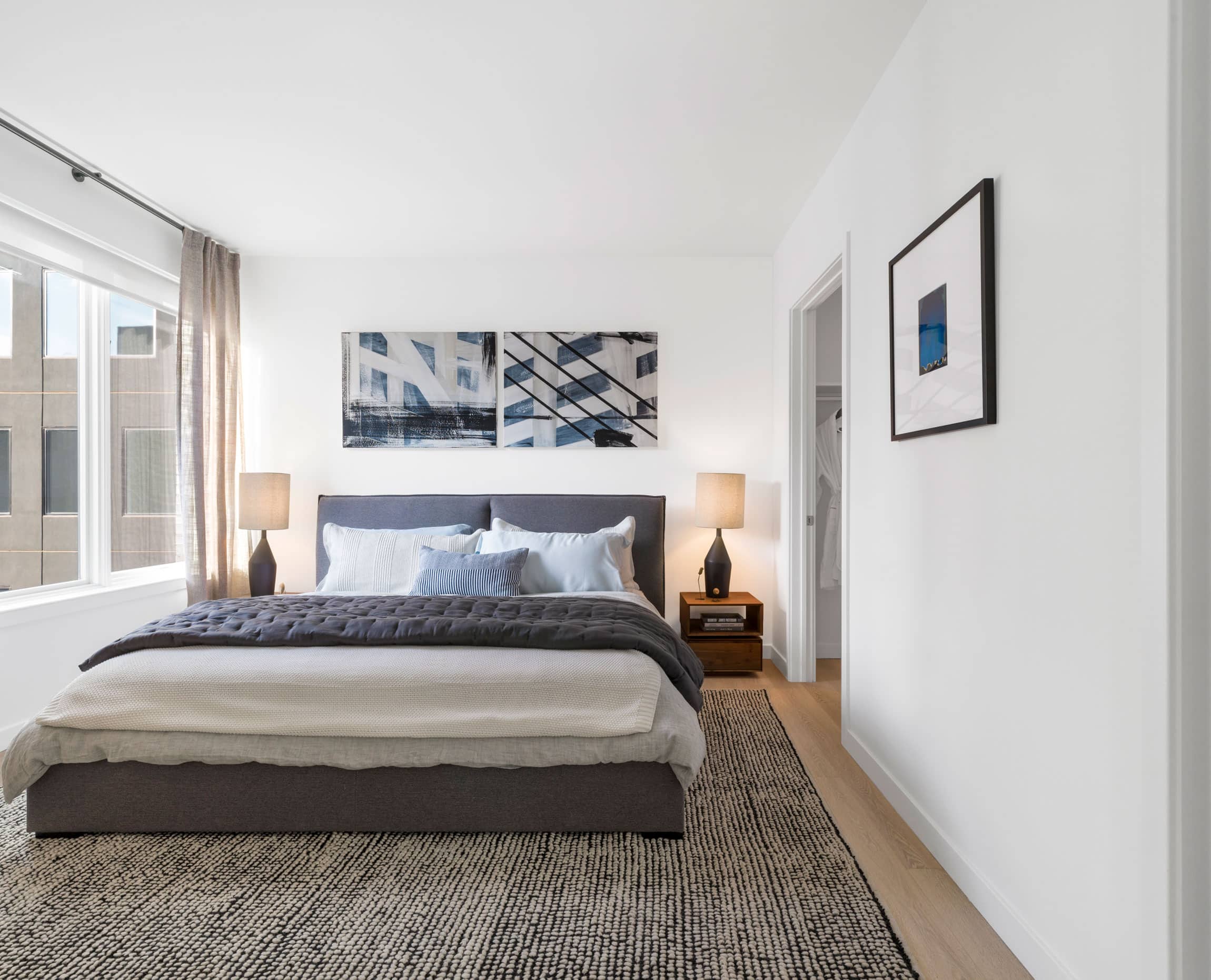 modern apartment bedroom with bed with gray comforter, nightstand, lamps and large window