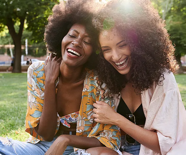 Two joyful friends laughing, enjoying a sunny day in the park with lush green surroundings.