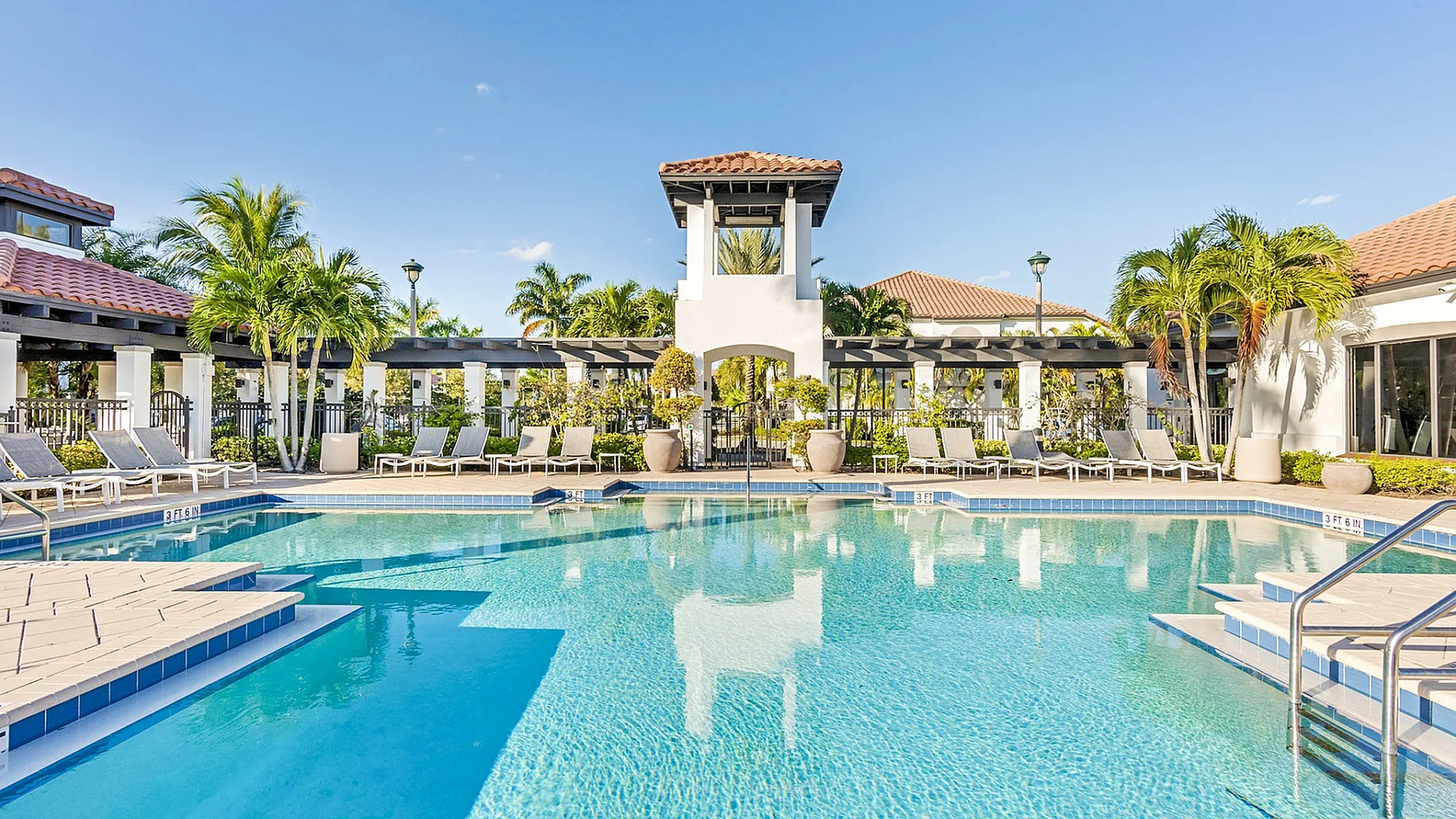 Luxurious resort-style swimming pool surrounded by sun loungers, tropical palm trees, and elegant architecture under a clear blue sky.