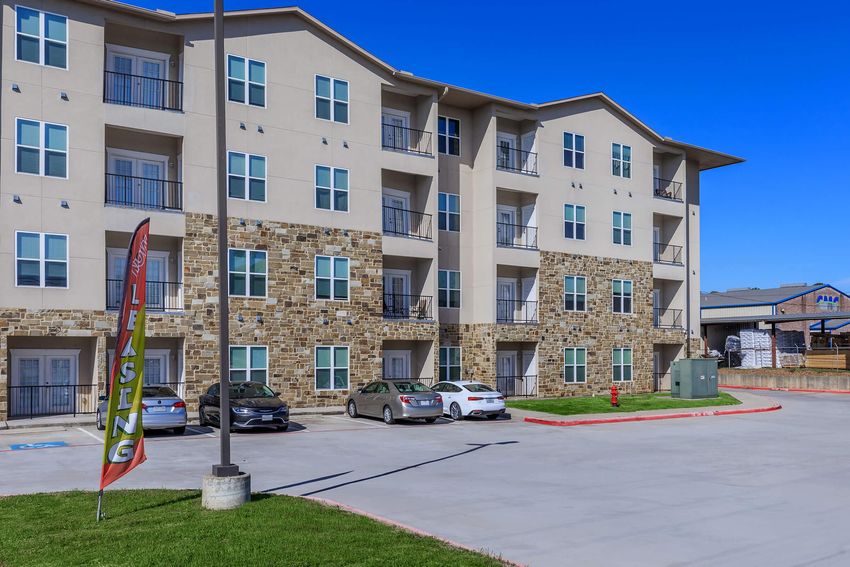 A multi-story apartment building with a beige exterior and stone accents. There are several balconies, windows, and parked cars in the foreground. A colorful leasing sign is prominently displayed, and the parking area is paved with a clear blue sky above.