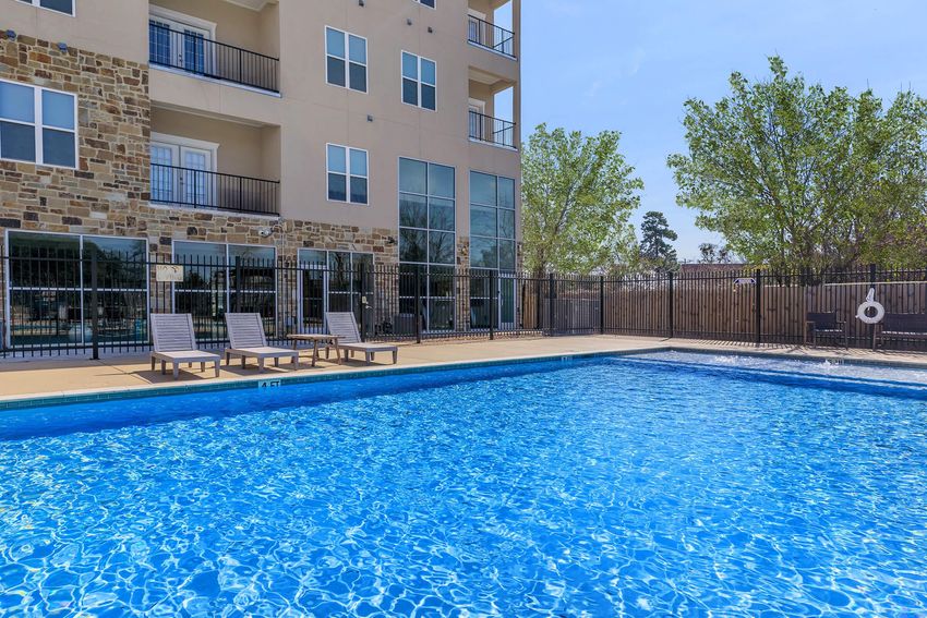 A clear blue swimming pool surrounded by lounge chairs, set in a well-maintained area next to a modern apartment building with large windows. Lush green trees frame the scene, and a safety buoy is visible near the pool's edge, creating a serene, inviting atmosphere for relaxation.