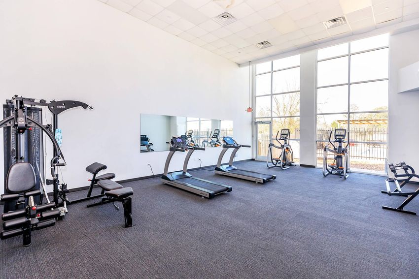 A spacious gym area with exercise equipment, including a multi-gym machine, treadmills, and a mirror. The room has large windows allowing natural light, with a minimalist design featuring a bright, neutral color scheme and a clean, modern aesthetic.