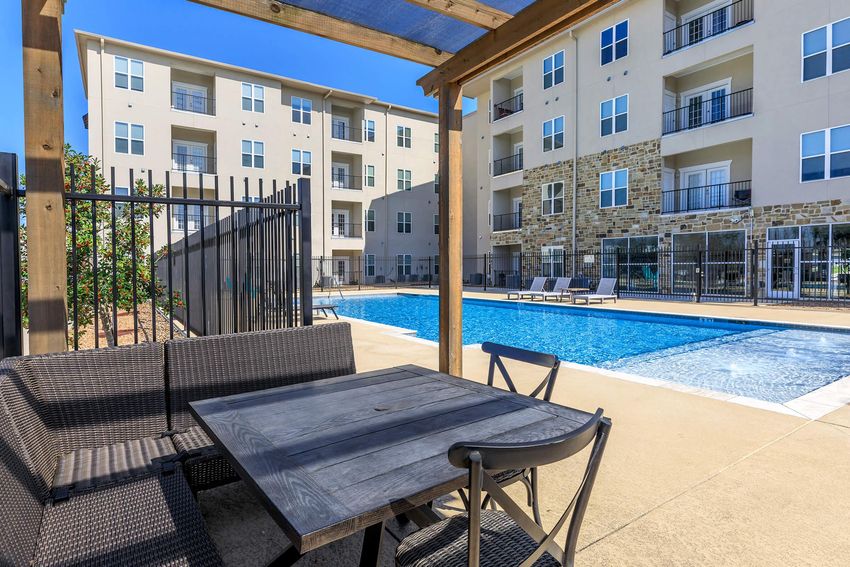 A view of a swimming pool surrounded by modern apartment buildings. There is a shaded seating area with a dark wooden table and chairs in the foreground. The pool features lounge chairs and is enclosed by a black fence. Bright blue skies and well-maintained landscaping complete the scene.