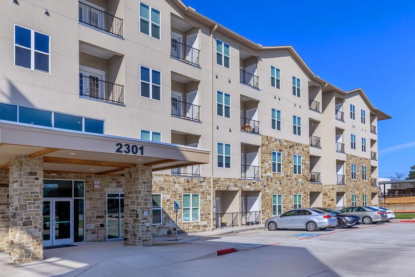 Modern multi-story apartment building with a stone and stucco exterior. The entrance features glass doors and a covered entryway. Several vehicles are parked in front, and the building has multiple balconies. Bright blue sky visible in the background.